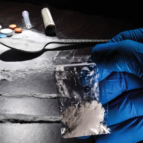 Gloved hand holding bag of white powder with drug paraphernalia in background