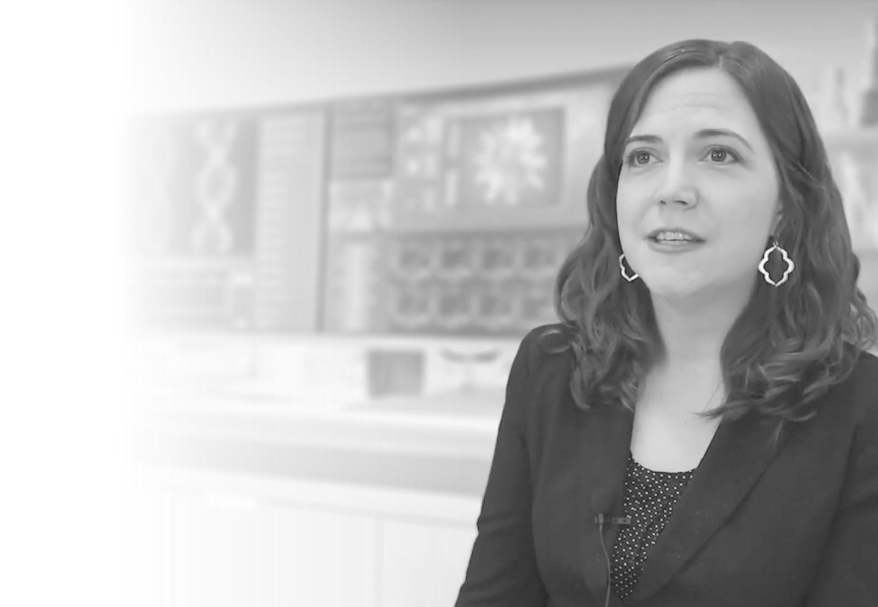 A white woman in a suit jacket faces the camera in front of a wall of diagrams