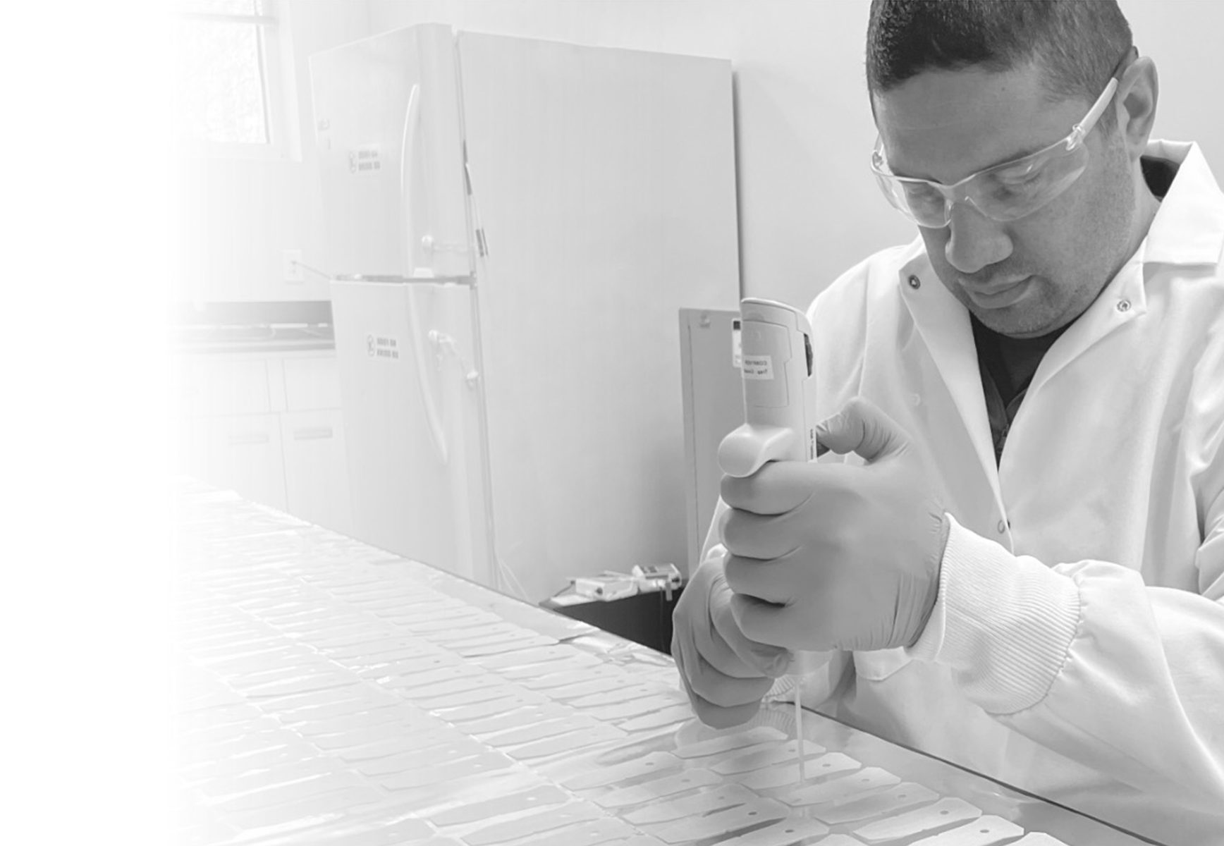 A man wearing a lab coat and goggles uses a pipette to apply samples to a number of test strips