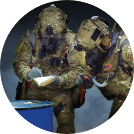 Two soldiers in full protective gear assessing a drum of unknown material during a training exercise.