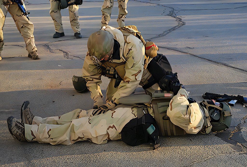 Photo of a training exercise with personnel in protective clothing. One person is lying on the ground simulating an injury while another is demonstrating how to assist him.