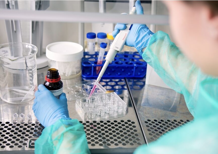 Technician in a lab pipetting liquid from a bottle to a vial under a safety hood.